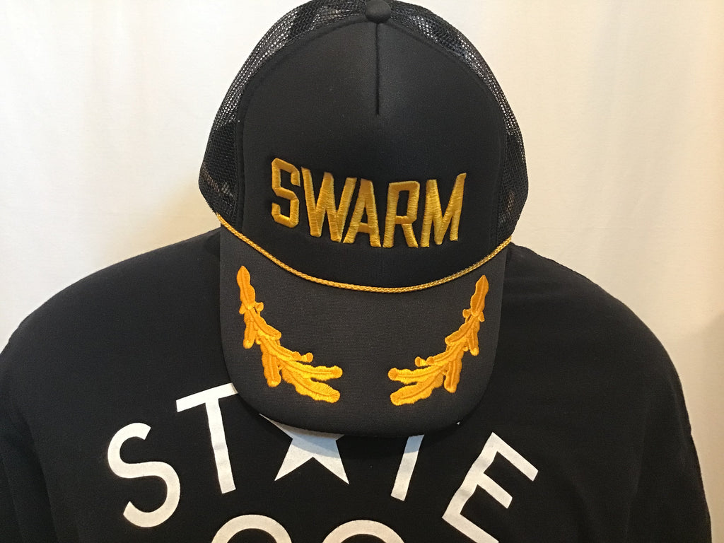 black foam front trucker hat with SWARM in gold embroidery a matching gold braid and gold oak leaves on the front hanging on mannequin