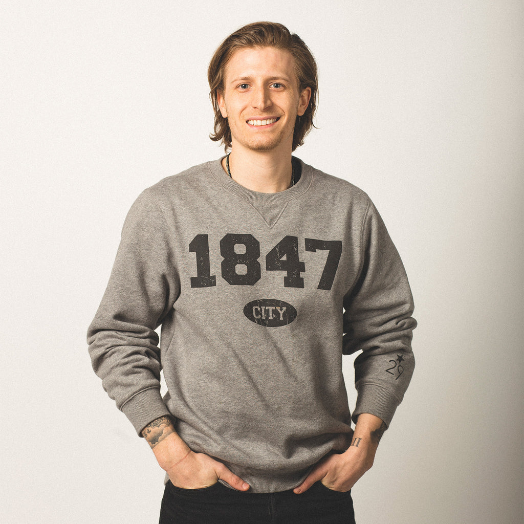 male model caucasian with brown chin length hair wearing crewneck vintage grey sweatshirt featuring bold black lettering of 1847 screen printed on front city in small bubble underneath wearing size medium