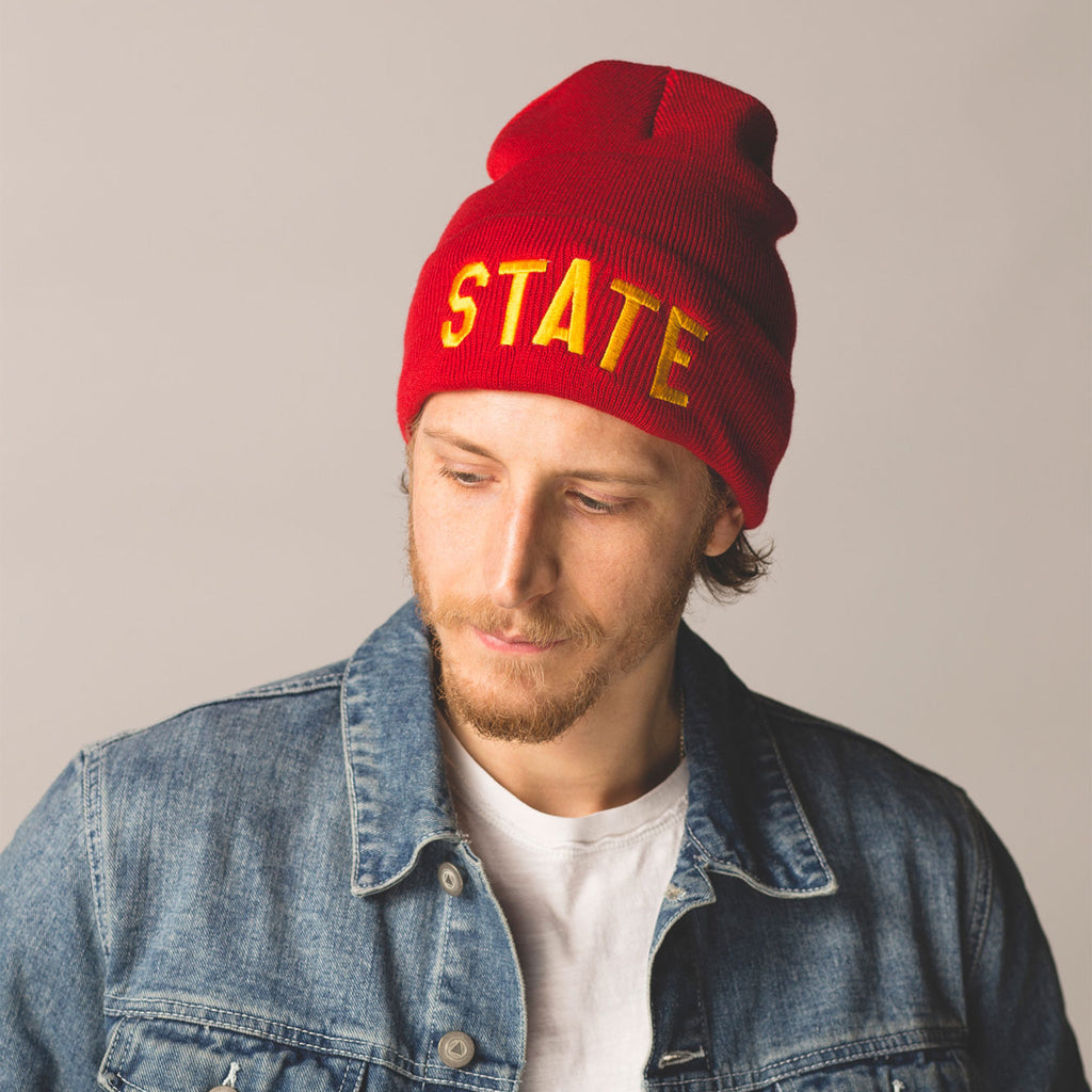 male model with short beard and mustache wearing red beanie with folded cuff state embroidered in gold lettering