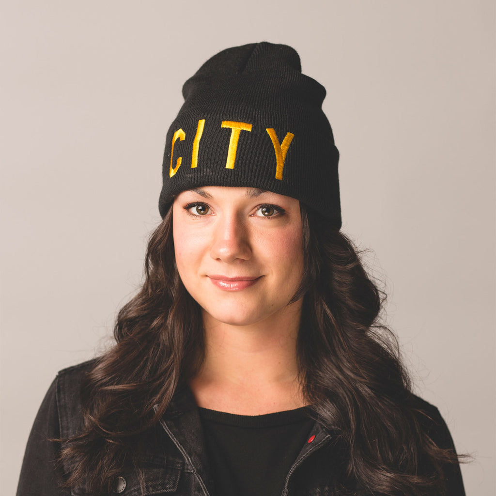 black haired model brown eyes smiling wearing black beanie with folding cuff city in gold bold lettering