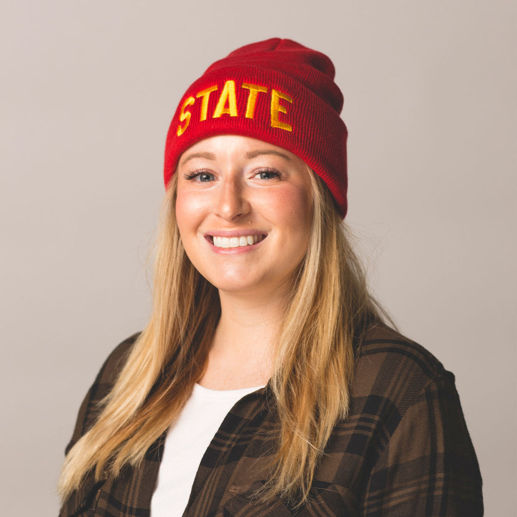 blonde long haired model smiling wearing red beanie with folded cuff state embroidered on front in gold