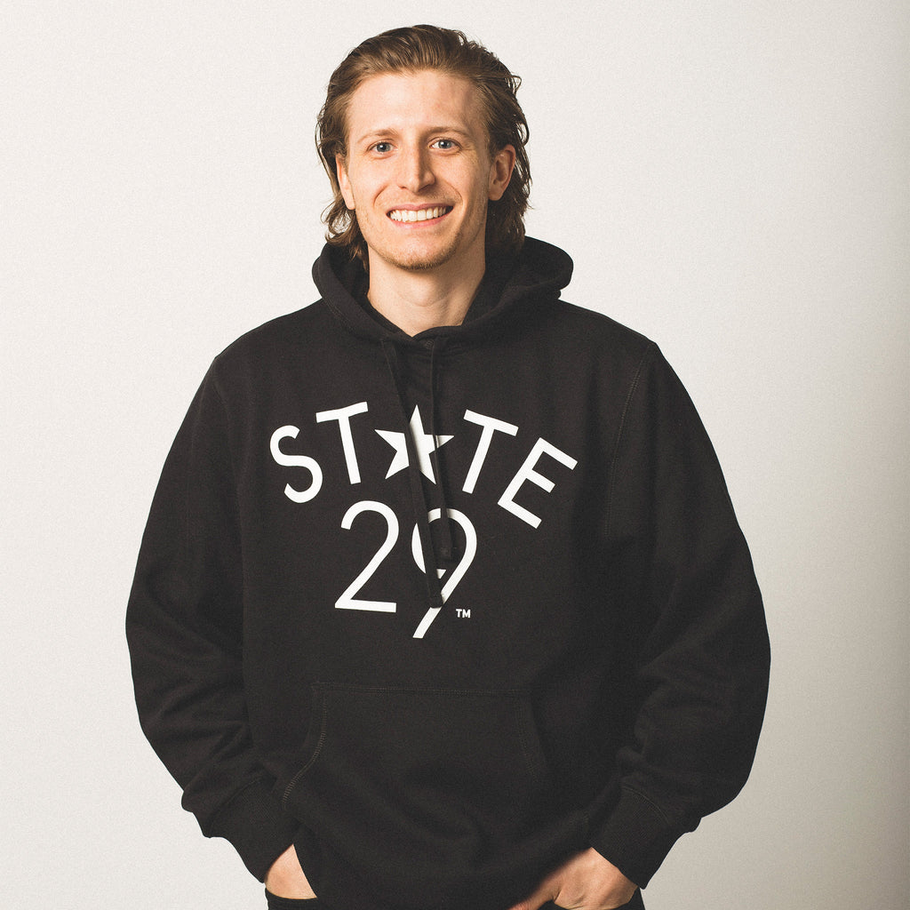 male model caucasian with brown chin length hair wearing pullover hooded black sweatshirt featuring white 29th State Apparel logo trademarked on front above pouch