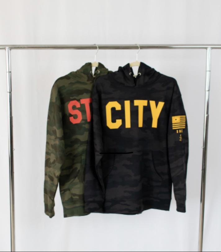 two hooded sweatshirts hanging from a silver rack a black camoflauge military inspired sweatshirts hangs first city in gold on front in bold details also in gold on left sleeve