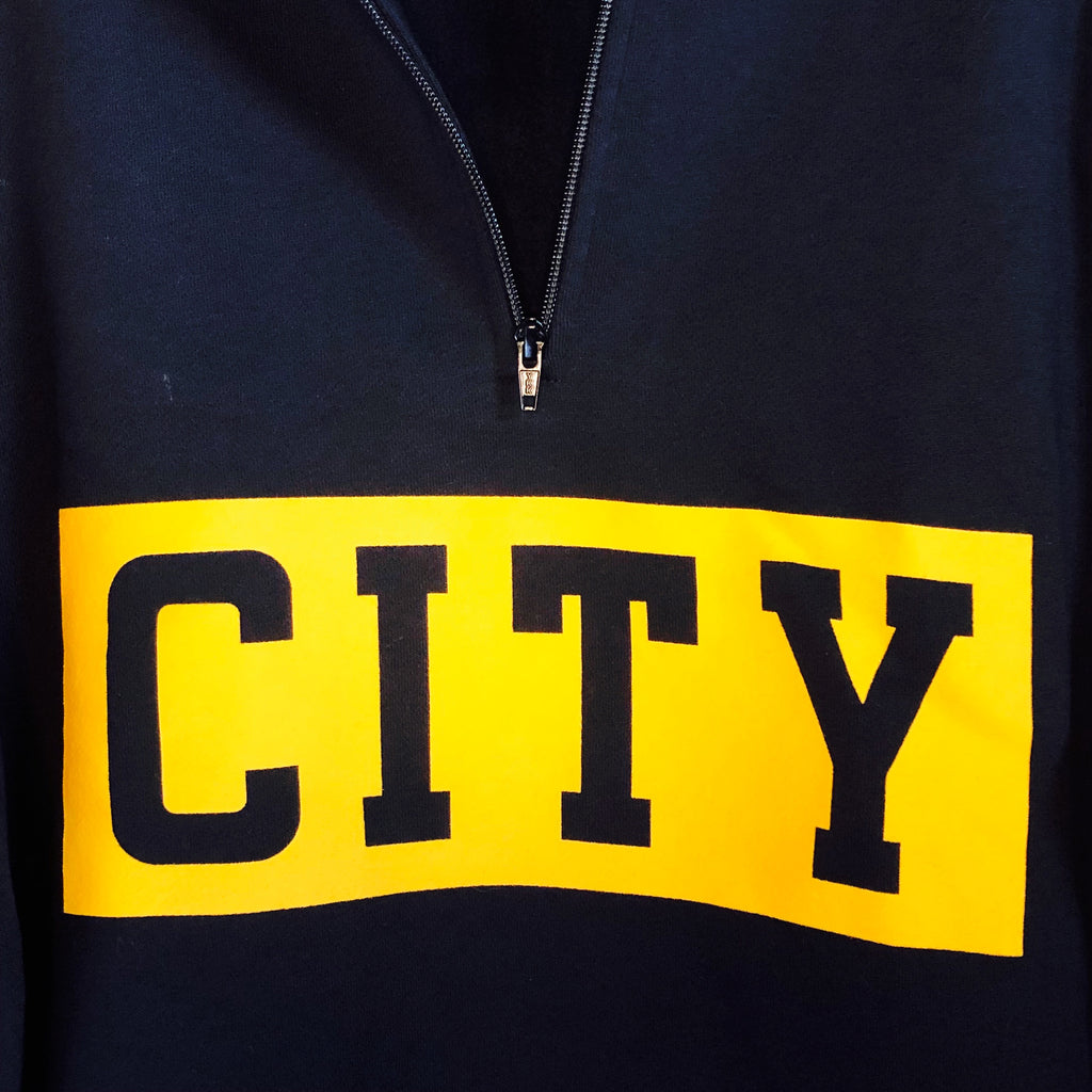close up of black 1/4-zip with bold gold stripe screen printed across the front and "city" cut out from screen print