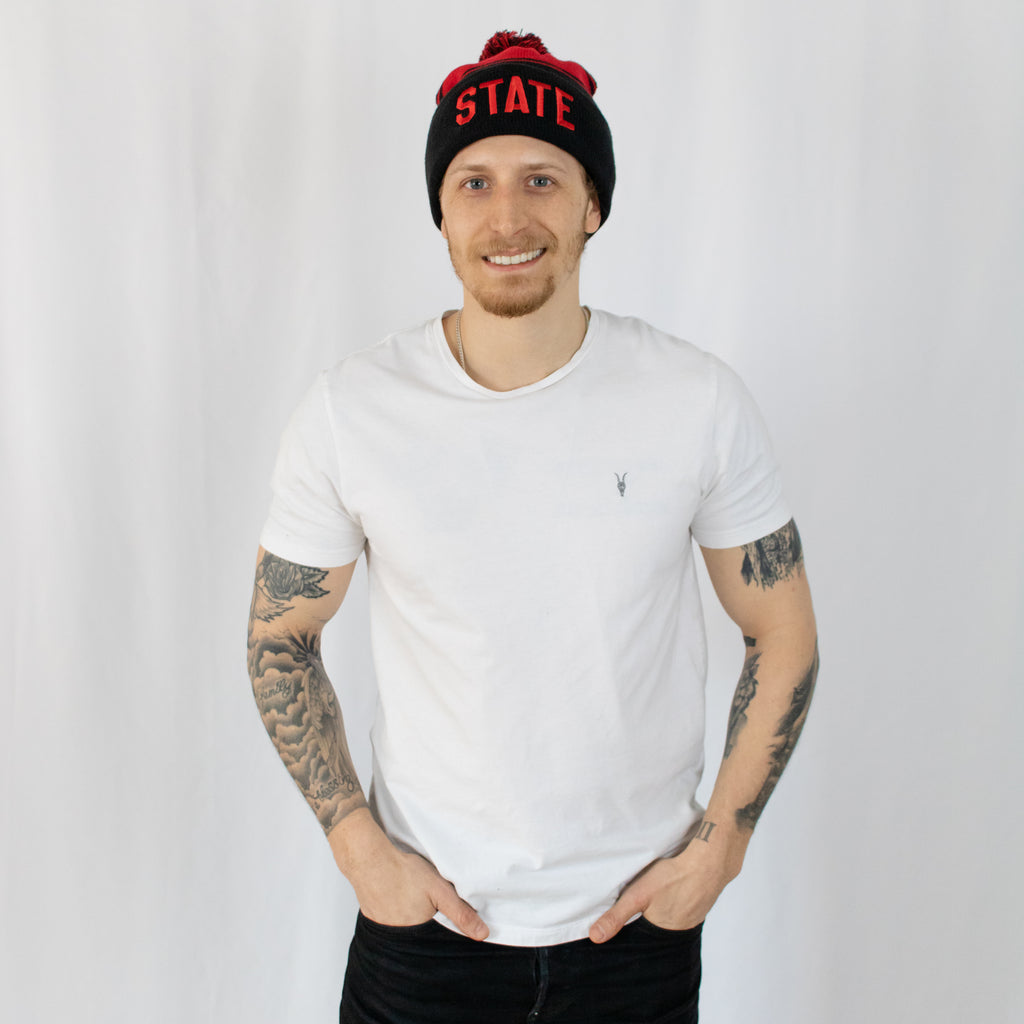 tattooed young man smiling in a white tee shirt wearing a two-tone black and red striped beanie state in red on front fold