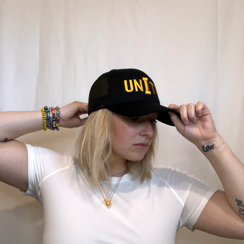 young women with blonde hair reaching for the front bill and back of an all black hat on her head unity embroidered in gold on front