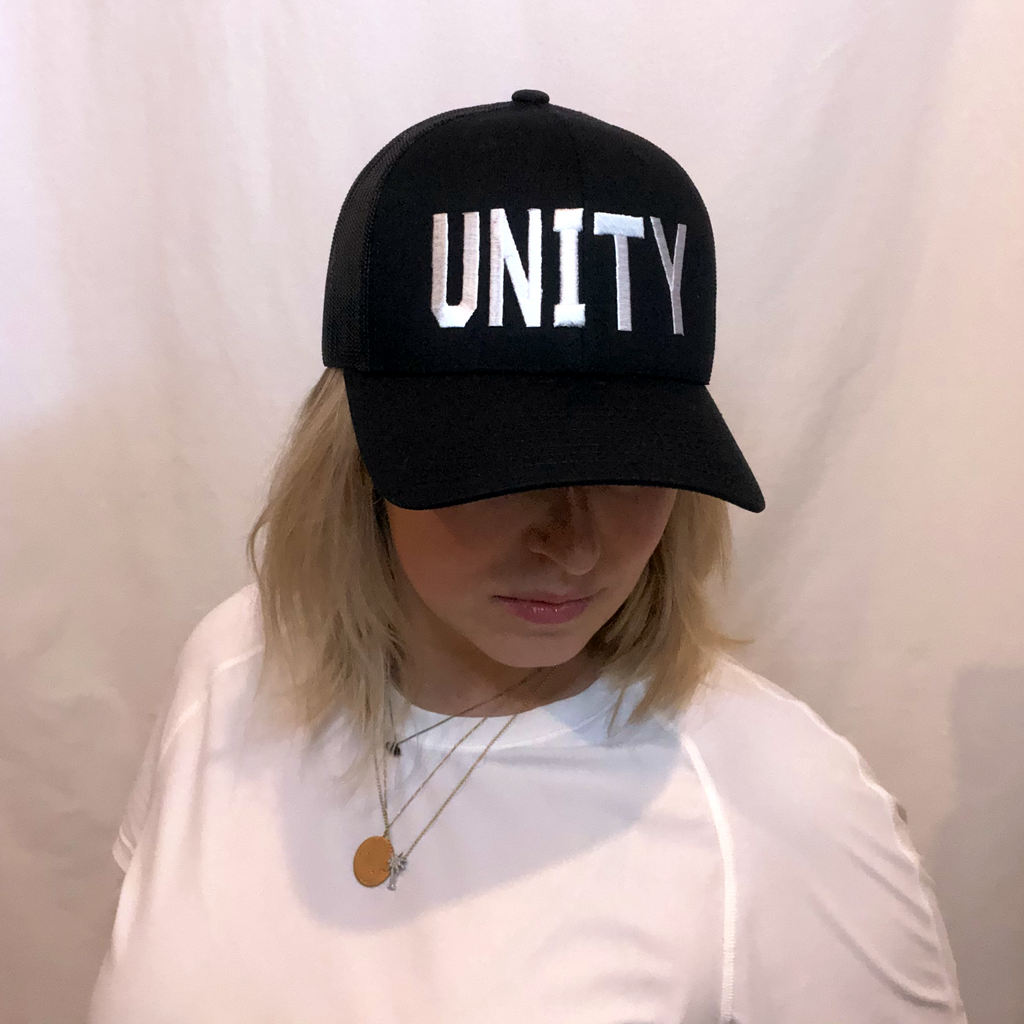 blonde haired girl in an all black trucker hat with unity in white embroidered on front