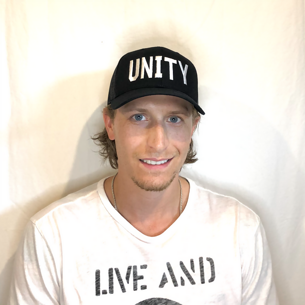smiling blonde young man wearing a black trucker hat with unity in white embroidered on front