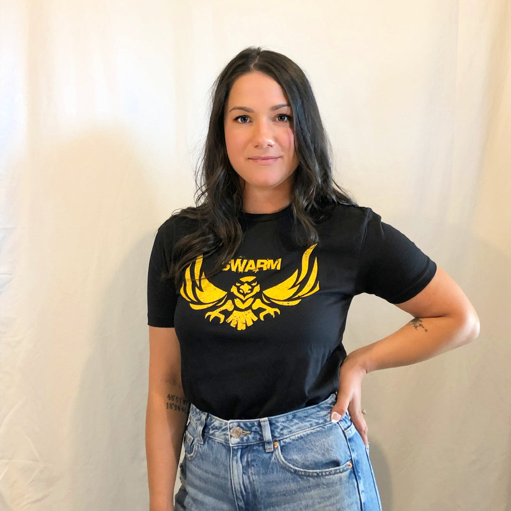 Young lady with long black hair and her hand on her hip in a pair of jeans and a black t-shirt with swarm in block lettering in gold above a hawk in gold