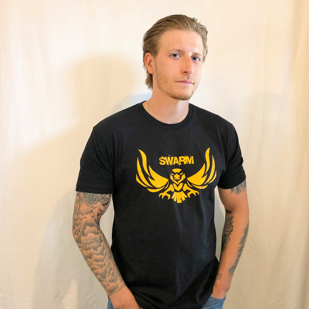 Young man with slicked back hair and a scruffy beard in a black t-shirt with swarm in block letters above a hawk in gold