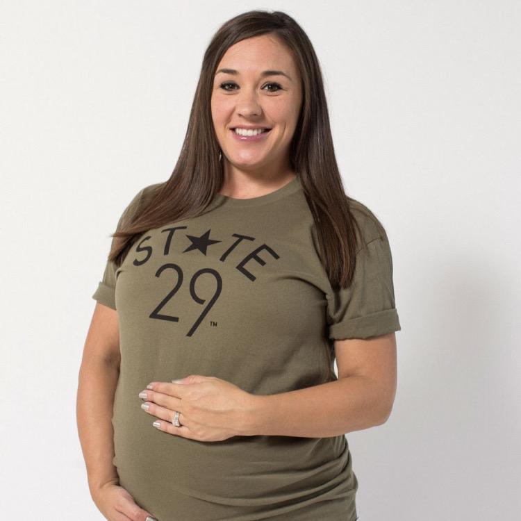 pregnant brunette model holding her bump wearing an olive green tee with the 29th state apparel logo on the front in black