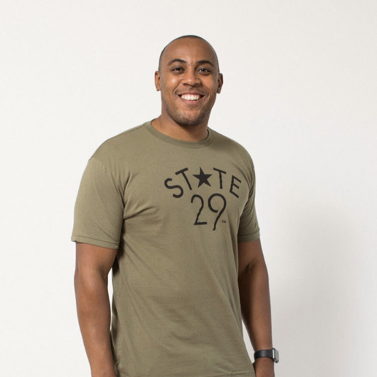 tall dark young man wearing an olive green tee with the 29th state apparel logo on the front in black