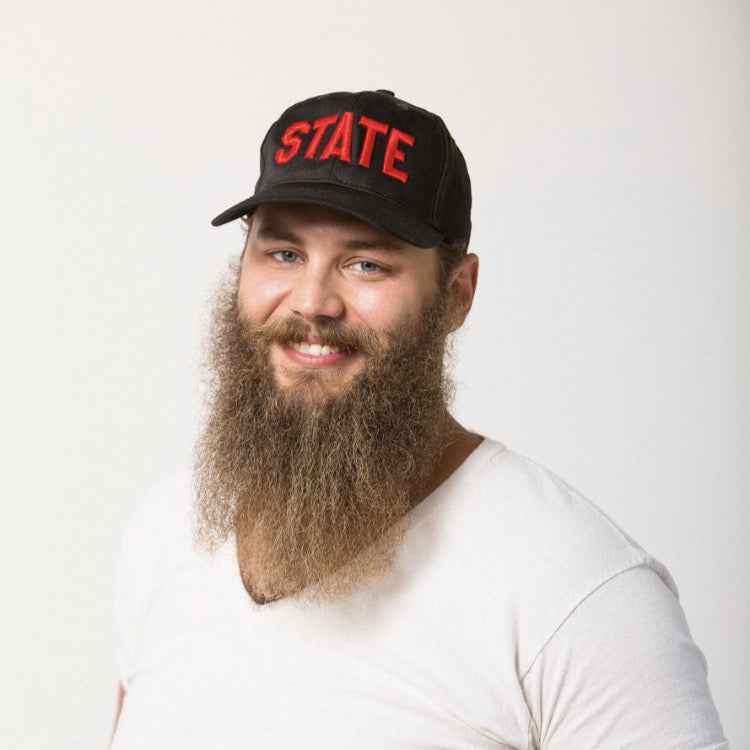 bearded male smiling in a black baseball hat with STATE embroidered in red on the front