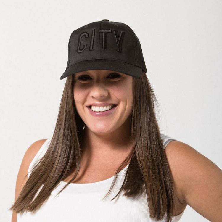 long dark haired woman smiling with hand on her hip wearing a black on black CITY baseball hat