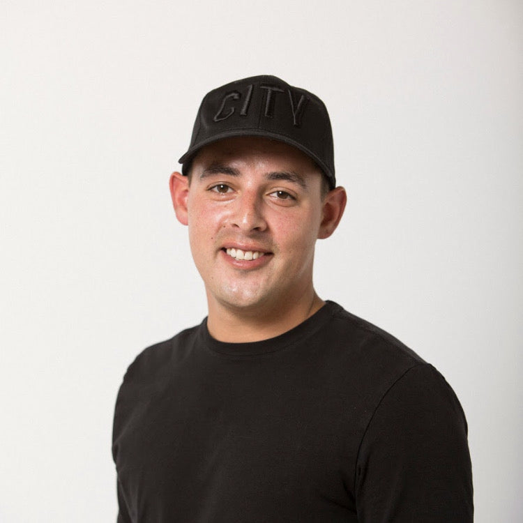dark haired young man in a black tee wearing a black on black CITY embroidered baseball hat