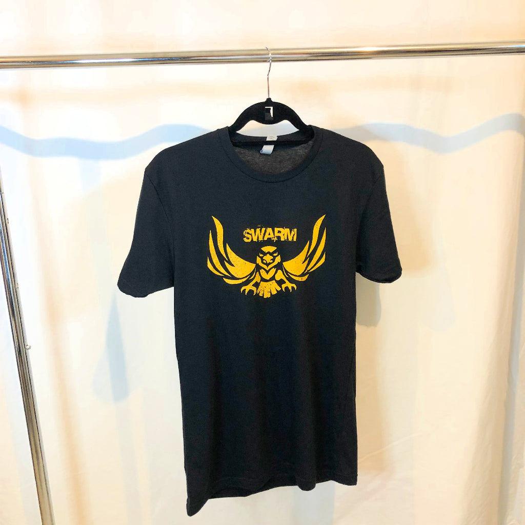 A black t-shirt hanging from a hanger on a rack the t-shirt has swarm in block lettering above a hawk all the details are in gold